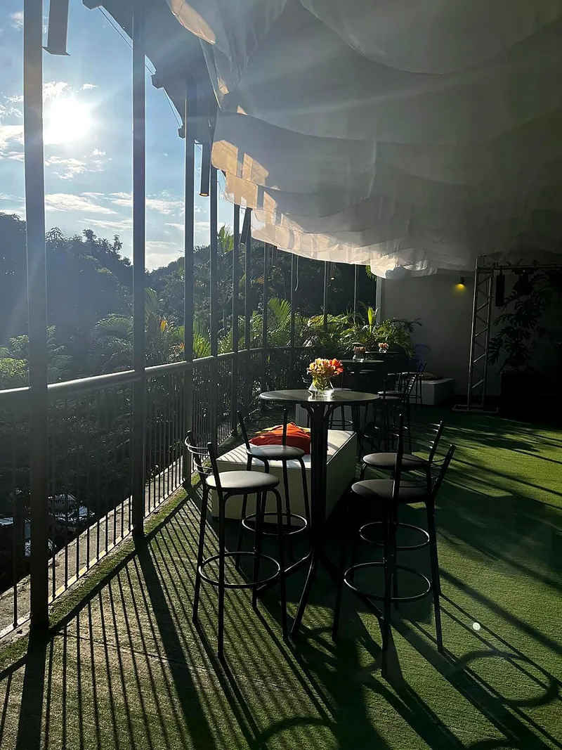 Varanda com guarda-corpo, mesas bistrô e sol entre as árvores