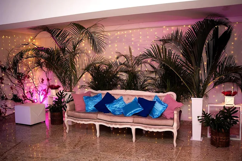 Lounge com sofá clássico, almofadas azuis e palmeiras iluminadas