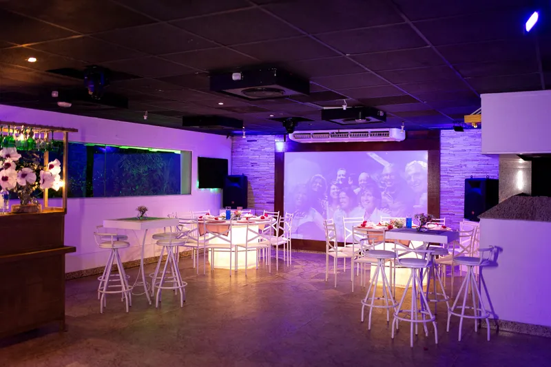 Salão com bar, telão e mesas bistrô iluminadas